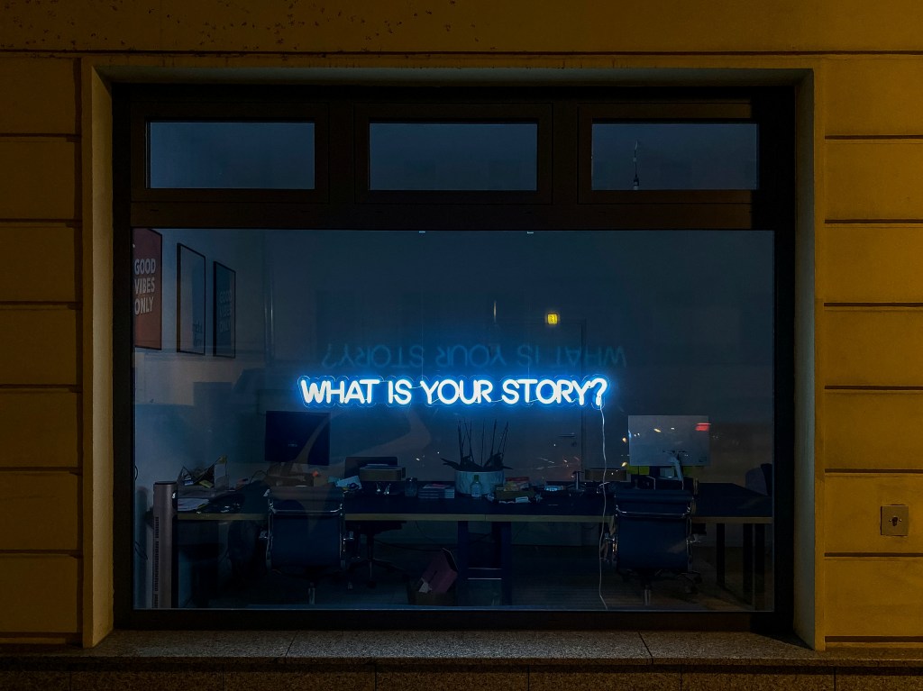 Photo by Etienne Girardet on Unsplash image of a neon light asking what is your story in front of an office window