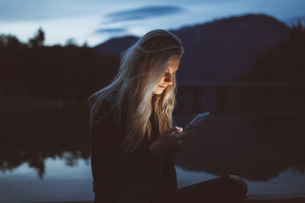 photograph of female subject looking at a lighted mobile device outdoors near a body of water after dark